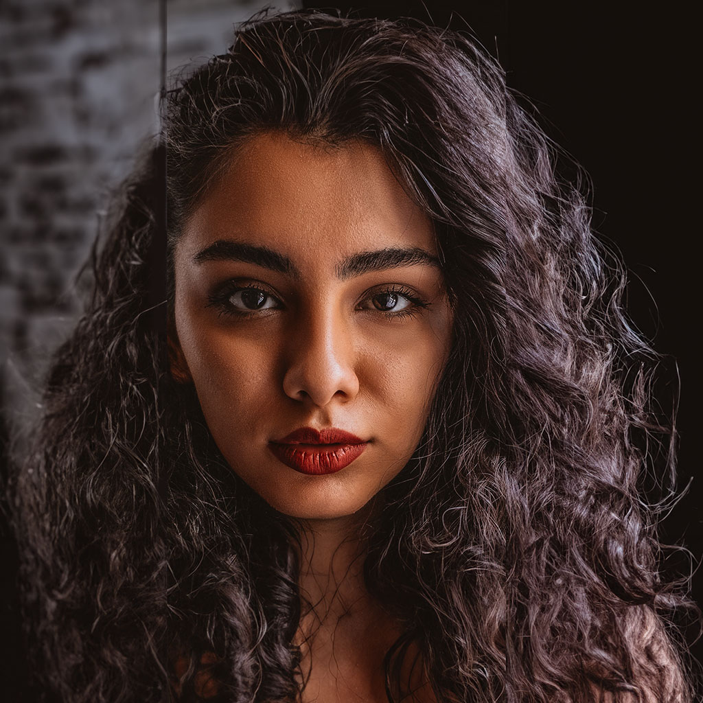 A person with long, curly hair and red lipstick stands in front of a brick wall, looking directly at the camera with a neutral expression.