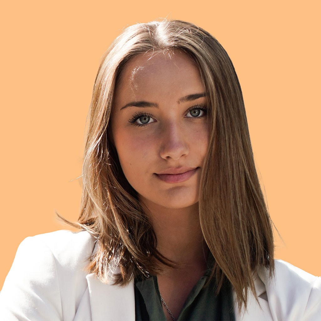 A young woman with long, straight brown hair is facing the camera against a solid orange background, wearing a white blazer over a dark top, exuding a professional and confident appearance.