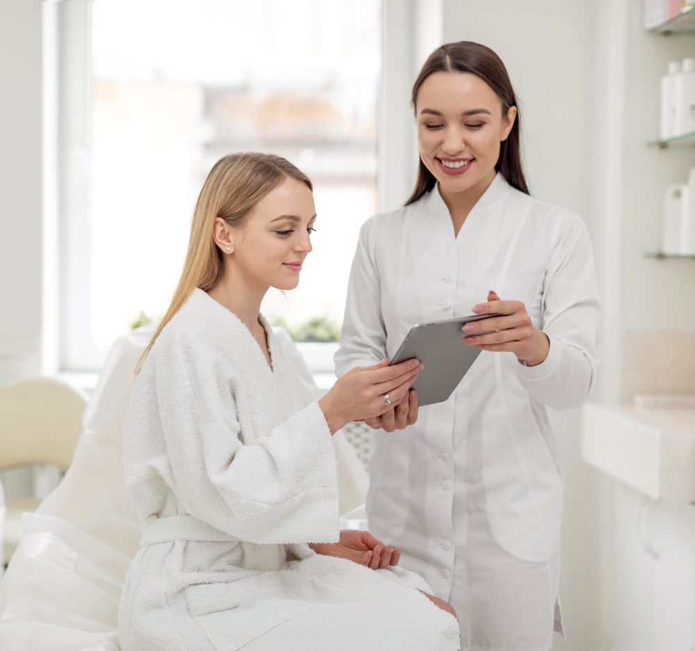 Two women in a spa setting, one in a white bathrobe and the other in a white uniform, smile as they look at a tablet, surrounded by calming, neutral-toned decor.